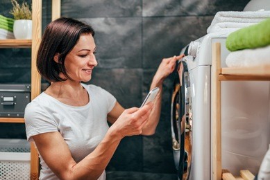 woman wearing white shirt using smart phone to control washing machine at laundry room