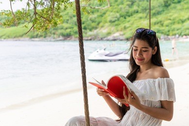 asian girl in white dress smiling happily by the sea for summer, traveling on vacation. read books on the beach swings on summer vacation.