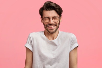 people and positive emotions concept. delighted glad bearded male with stylish hairdo, dark beard, keeps eyes shut, smiles joyfully, dressed in casual white t shirt, isolated over pink background
