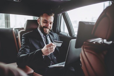 young handsome businessman is sitting in luxury car. serious bearded man in suit is working with laptop with credit card in hands.