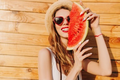 stunning blonde girl chilling in summer day and eating watermelon. close-up portrait of laughing european lady in boater and sunglasses fooling around on wooden background.