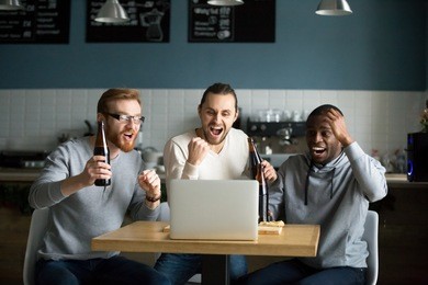 multiracial men drinking beer celebrating victory watching online football game match on laptop in pub restaurant, diverse friends supporting team excited with goal result, sport betting win concept