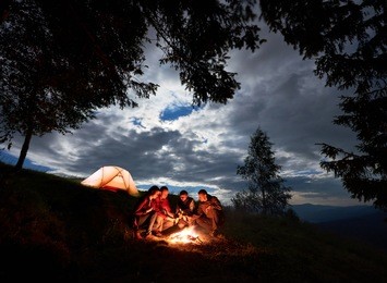 night camping in the mountains. group of friends hikers having a rest around campfire with beer, enjoying a holiday near glowing orange tent and forest under the cloudy sky at dawn