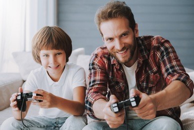 cheerful father with game controller plays video game with small son sitting on couch. parent spends time with child.