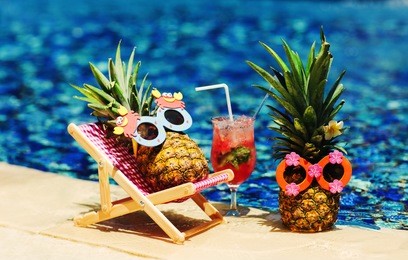 couple of attractive pineapples in cheerful sunglasses lying on sunchair near the turquoise swimming pool. relaxing, drinking fresh juice. tropical summer vacation concept. happy sunny day. sunbathing