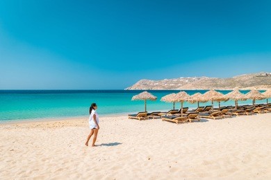 mykonos beach during summer with umbrella and luxury beach chairs beds, blue ocean with mountain at elia beach mikonos greece, young woman on the beach in bikini swim wear