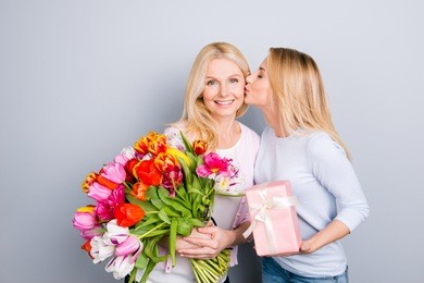 lovely girl kissing in cheek her mom prepare gift case in pink package with white bow, holding big bouquet of colorful tulips in hands enjoying  rest relax leisure isolated on grey background