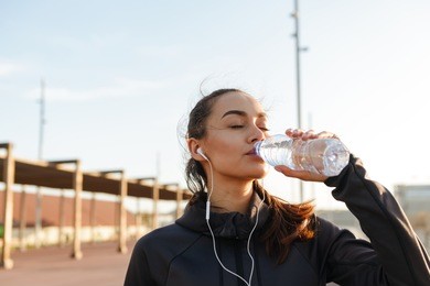 image of beautiful young asian sports woman listening music with earphones drinking water outdoors.