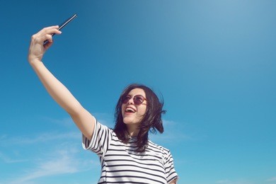 portrait of a cute asian teenager young woman stand against blue sky and cloud background. wearing sunglasses. take selfie and smile with smartphone. light effect added. travel summer hipster concept