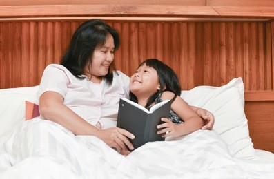 happy mother and daughter reading book on bed, portrait of cute girl listening to her mother telling an interesting story at home, family and education concept