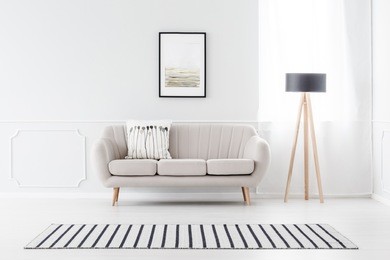 beige couch standing against white wall with molding and watercolor poster in cozy living room interior