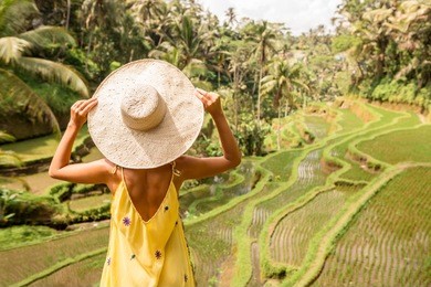 beautiful young lady in shine through dress touch straw hat. girl walk at typical asian hillside with rice farming, mountain shape green cascade rice field terraces paddies. ubud, bali, indonesia.