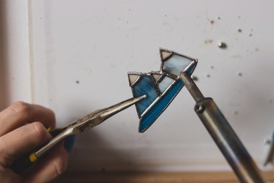 woman soldering stained window glass bijouterie creating handmade adornment in mask indoors. craftsman brazing making jewelry at home protection respirator mask. vitrage technique