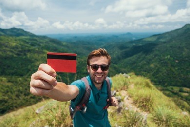 young smiling male showing empty credit card. he is standing on top of mountain and showing the outdoor beauty. concept of easy travelling