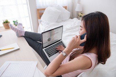 asian female freelancer talk on mobile and see business graph on laptop screen with rest leg on table in bedroom at home.work at home concept.work from home concept
