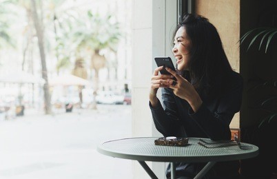 portrait of a cheerful asian woman looking at the street while sitting in a coffee shop with a mobile phone in her hands. business woman happy to read good news on a smartphone form business partners 