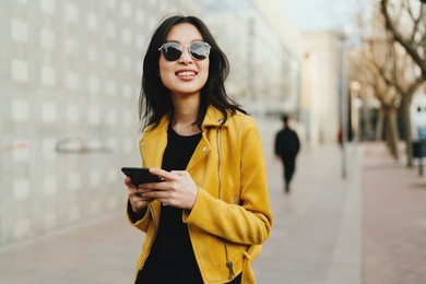 half length portrait of a beautiful smiling asian woman in a sunglasses with long dark hair wearing yellow leather jacket chatting online by a mobile phone while standing on blurred street background.