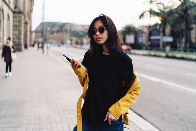 charming asian woman in sunglasses wearing blank black t-shirt and yellow leather jacket reading messages on a mobile phone while standing on a blurred city background. hipster girl holding smartphone