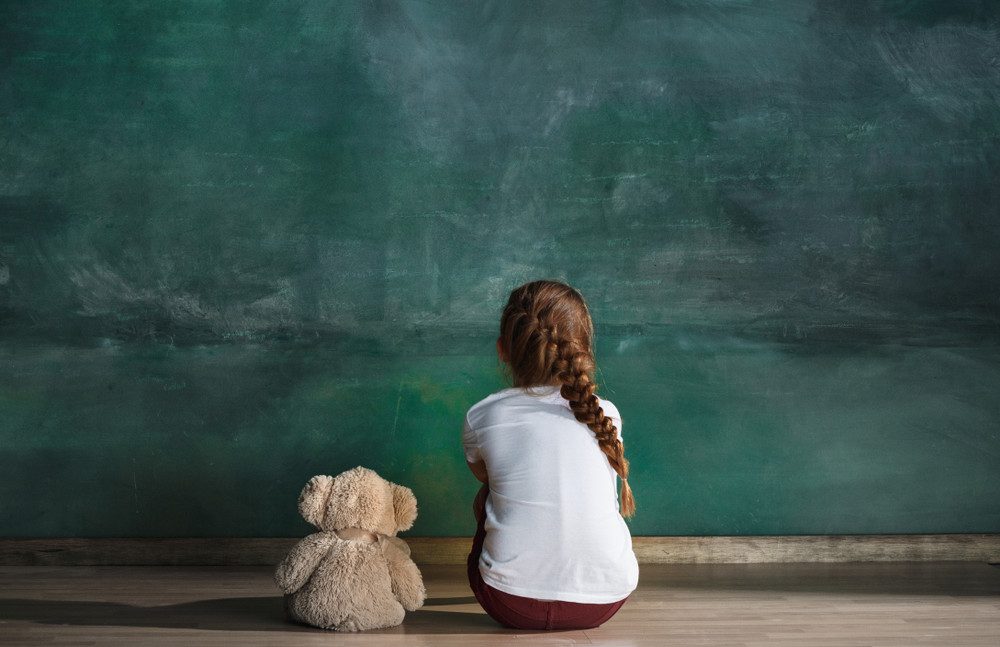 little autistic girl with teddy bear sitting on floor at empty room. autism concept. conceptual image with little caucasian model at studio. back view