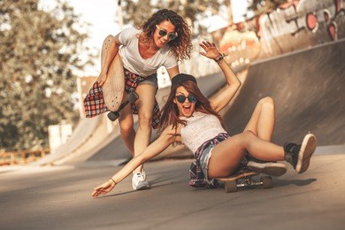 two female friends playing with skateboard at the skate park.one girl pushing other from behind.laughing and fun.