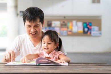 asian father and daughter are reading the storybook together and they are smiling with happy moment, concept of learning activity for kid in family lifestyle.