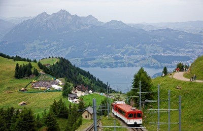 a sightseeing train travels on the cogwheel railway thru green grassy meadows on mt. rigi, with rugged pilatus peaks among alpine mountains opposite lake lucerne on a cloudy summer day in switzerland