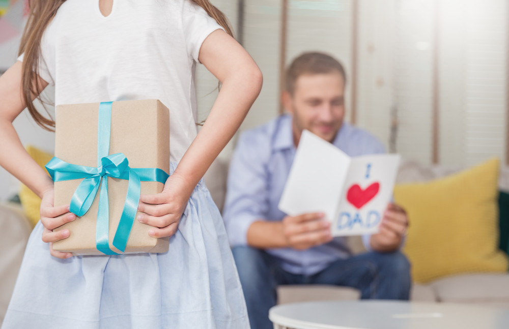 little caucasian girl daughter congratulate daddy hiding giving a surprise card and gift box. family holiday father and girl celebrate together. love lifestyle happy father’s day holiday party concept