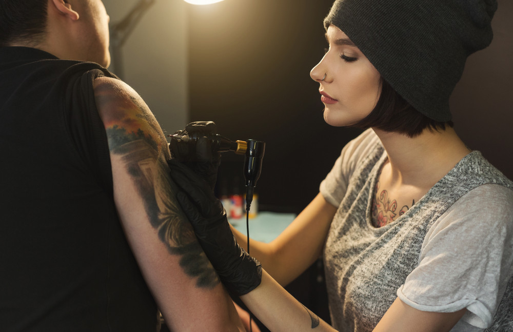 concentrated woman tattooist doing tattoo with tattoo machine on male shoulder. unusual female profession, copy space