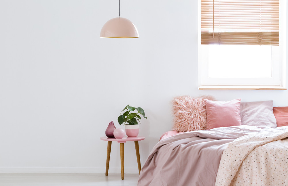 cozy, feminine bedroom with pink bed, decorative cushions and plant on a wooden stool standing against white, empty wall. real photo with a place for your furniture.