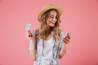portrait of a happy young woman in summer dress and straw hat showing plastic credit card while using mobile phone isolated over pink background