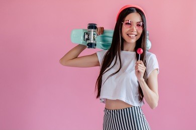 young asian teenage girl on pink background. stylish girl with lollipop and skateboard isolated.