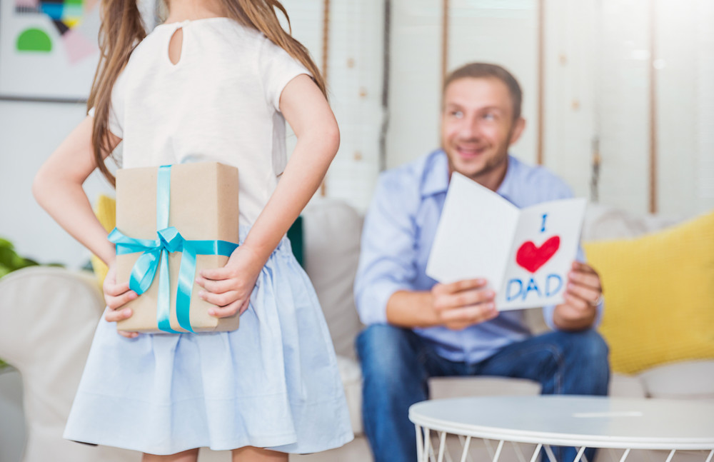 little caucasian girl daughter congratulate daddy hiding giving a surprise card and gift box. family holiday father and girl celebrate together. love lifestyle happy father’s day party concept