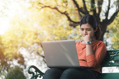 young asian woman working with her laptop on and thinking about her online business bench in the park outdoors on vacation time.