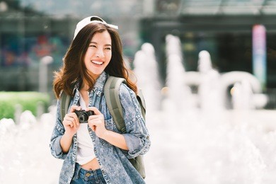 young beautiful asian backpack traveler woman using digital compact camera and smile, looking at copy space. journey trip lifestyle, world travel explorer or asia summer tourism concept