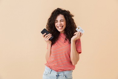 photo of cute pretty young curly woman isolated over beige background using mobile phone holding credit card.