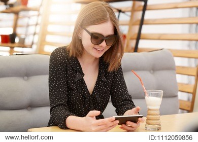 satisfied female in trendy shades and blouse, watches interesting video on mobile phone, connected to wireless internet at coffee shop, drinks milk shake, has joyful expression, has spare time