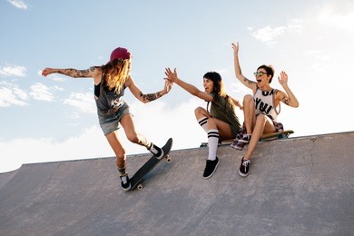 female skateboarder riding skateboard at skate park with friends sitting on ramp having fun. woman skater giving high five to female friend sitting on ramp during her routine.