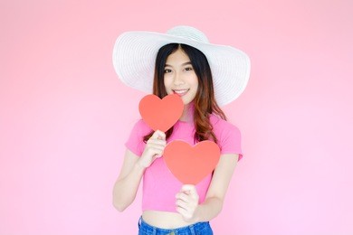 portrait beautiful asian woman wearing pink t-shirt and white hat on pink background, happy valentine day in love concept, model holding red heart shaped paper in hand