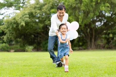 asian father and daughter are playing the balloons and they running and laughing together with fully happy moment in the park, concept of outdoor activity in family lifestyle.