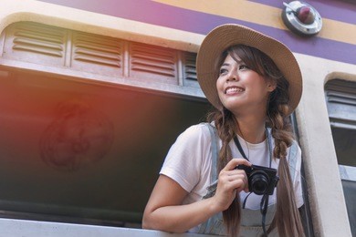attractive cheerful asian young woman wearing hat from window train holding camera in hand. young girl leans out of the train window for looking interest place or tourist location. 