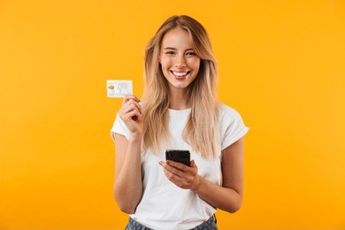 portrait of a happy young blonde girl showing plastic credit card while holding mobile phone isolated over yellow background