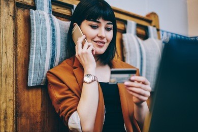 positive woman looking at number on credit card and confirm purchase via telephone call to customer service, smiling hipster girl making payment via smartphone conversation while resting indoors