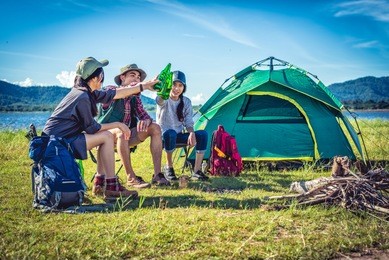 group of young asian friends enjoy picnic and party at lake with camping backpack and chair. young people toasting and cheering bottles of beer. people and lifestyles concept. outdoor background theme