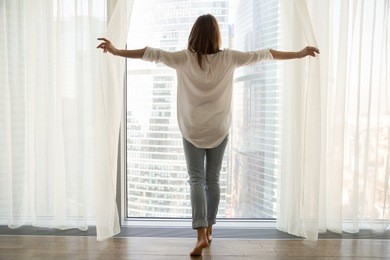 rear view at rich woman standing looking out of full-length window of luxury modern apartment or hotel room opening curtains in the morning enjoying sunlight and city skyscrapers view feeling happy