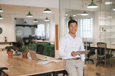 portrait of a smiling young asian businessman leaning on his desk in a large modern office working online with a digital tablet