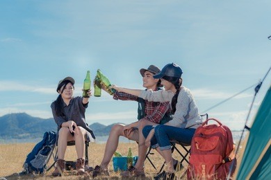 camping group of friends asian people together, teenagers travel with backpack,tent clink beers bottle relaxing on vacation time holiday sitting on chairs near river and mountain view.