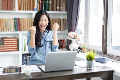 yes! happy excited asian woman at home with raised hands,enjoying time, beautiful young smiling girl working on laptop , celebrating the success business