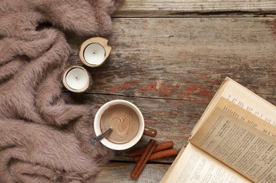 warm soft blanket, cup of hot chocolate, candles, cinnamon sticks and open book on old rustic wood background. cozy still life. top view point.