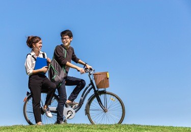young couple asian students ride a bicycle with textbook on grass hill in campus in university with copy space of clear blue sky background, in a concept education, love, sport, study, relax, friend 