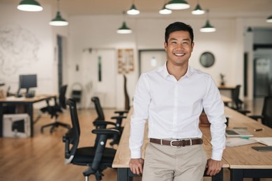 smiling young asian businessman leaning against a table while working alone in a large modern office